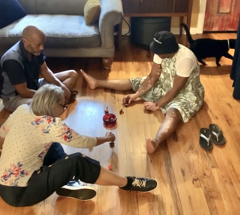 Three adults sitting on the floor of a living room playing a game of jacks together
