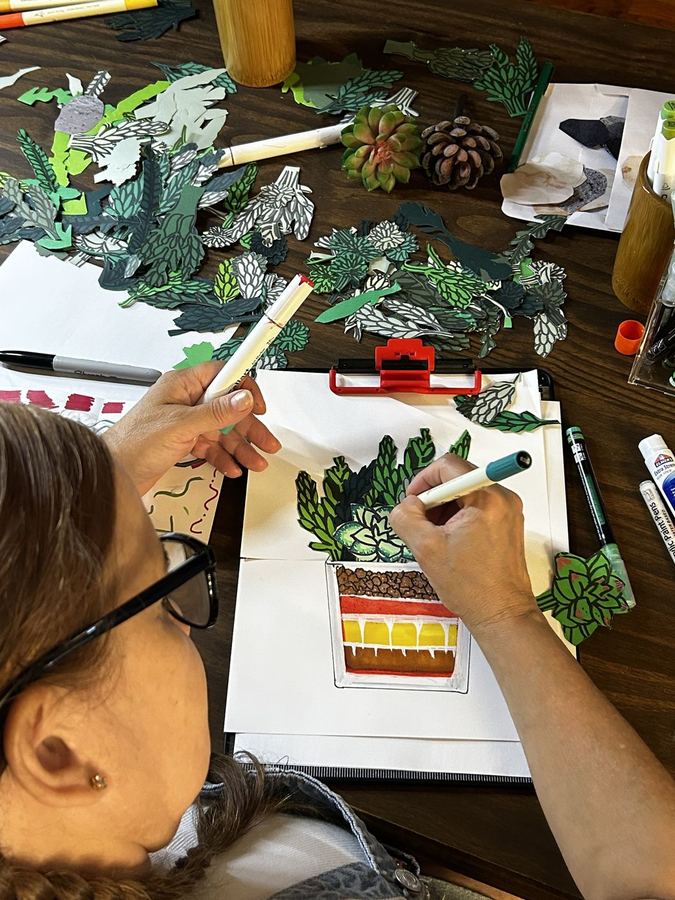 Close-up of a hand drawing a layered terrarium in a sketchbook surrounded by cut paper leaves