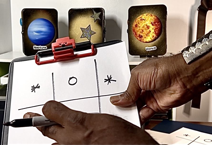 A hand drawing a tic-tac-toe grid beside star and planet cards